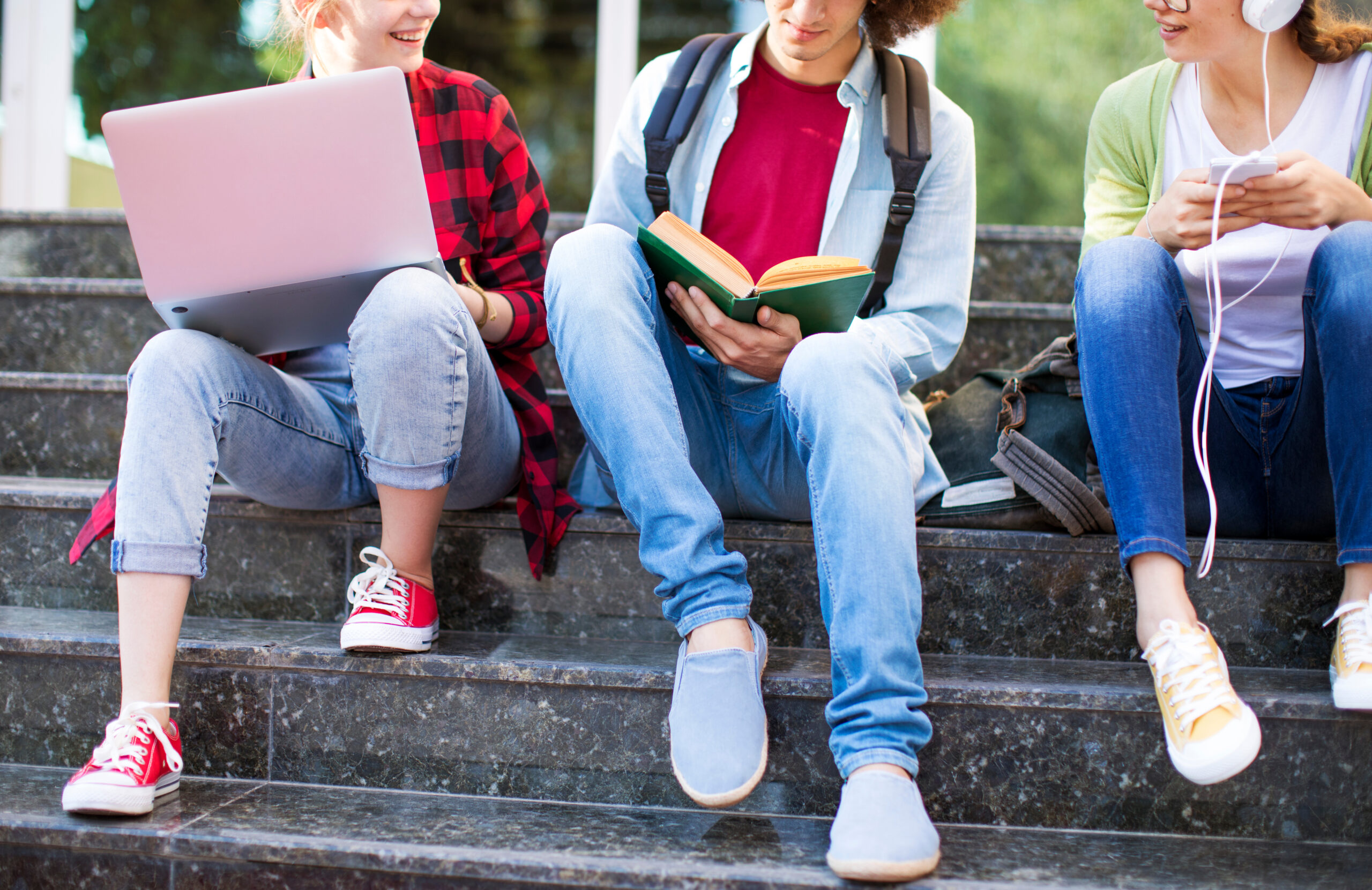 Three teenagers sitting on steps while one holds a computer one holds a book and one holds a phone connected to headphones.